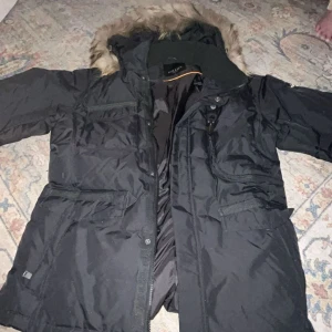 Svart vinterjacka från Hollies - Säljer en svart vinterjacka från Hollies i storlek 54. Jackan har en mysig äkta pälsfodrad huva och flera praktiska fickor. Huvan och pälsen går att ta av, var för sig. Så varm och go.