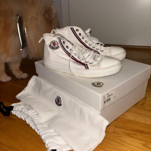 Vita sneakers från Moncler - Säljer ett par vita Moncler sneakers i bra skick. Köpta på nk i stockholm för 3000kr. De har en klassisk design med snörning och Moncler-logga på sidan. Skorna har röda och blå ränder längs sidorna som ger en snygg kontrast. Storlek 35. Påsen ingår skriv om ni vill ha kartong oxå.
