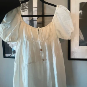 Klänning - Säljer en superfin vit klänning med puffiga kortärmar och en söt knytning framtill från Zara. Perfekt för sommarens alla tillfällen! Klänningen har en luftig passform, och är gjord i ett lätt material som känns skönt. En superbra studentklänning!! Passar perfekt till både stundeten och andra sommarfester. Endast testad. 