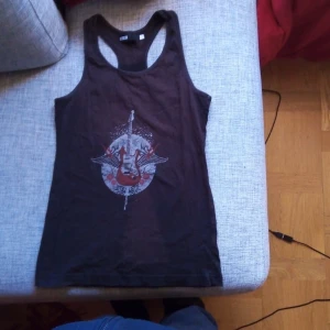 Svart linne med gitarrtryck från DSW - Säljer ett svart linne från DSW i storlek M. Det har ett coolt tryck med en elgitarr och vingar i rött och vitt på framsidan. Perfekt för sommarens festivaler eller en avslappnad dag. Linnet är i bra skick och redo för nya äventyr!