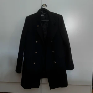 Svart kappa från Zara - Säljer en elegant svart kappa från Zara i storlek M. Den har en klassisk design med dubbelknäppning och långa ärmar. Perfekt för höst och vinter, och passar både till vardags och fest. Kappan är i mycket bra skick och redo för en ny ägare!