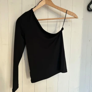 Svart oneshoulder topp - Snygg svart oneshoulder topp med en lång ärm och en smal axelrem på andra sidan. Perfekt för en utekväll eller fest. Materialet ser ut att vara mjukt och stretchigt för en bekväm passform.