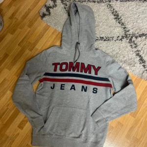 Grå hoodie från Tommy Jeans - Säljer en snygg grå hoodie från Tommy Jeans i storlek S. Den har en klassisk design med röd och blå text på bröstet och en bekväm huva med dragsko. Perfekt för en avslappnad stil och passar till alla årstider. Skön och stilren, ett måste i garderoben!