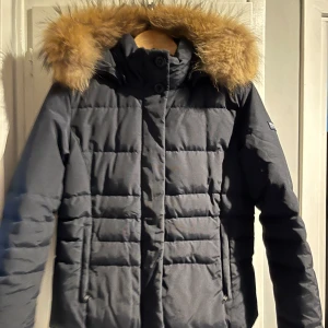 Mörkblå dunjacka från Woolrich - Säljer en snygg mörkblå dunjacka från Woolrich i storlek M. Jackan har en fluffig pälsfodrad huva och är perfekt för kalla vinterdagar. Den är i mycket bra skick och har en stilren design med dragkedja och knappar framtill.