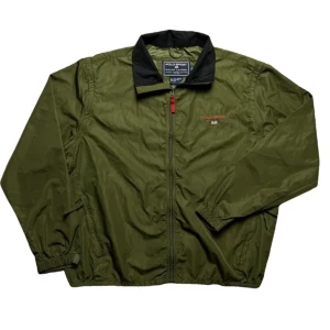 Grön vindjacka från Polo Sport - Säljer en grön vindjacka från Polo Sport, perfekt för höst och vår. Jackan är i storlek L och har en dragkedja framtill. Tillverkad i 100% polyester, vilket gör den lätt och bekväm. Den har en klassisk stil med en svart krage och Polo Sport-loggan broderad på bröstet. Passar både till vardags och lättare utomhusaktiviteter.