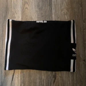 Svart tubtopp från Adidas - Säljer en svart tubtopp från Adidas med de klassiska tre ränderna i vitt. Toppen är i storlek S och har en stilren design med Adidas-loggan tydligt synlig. Perfekt för vardagsbruk. Nypris 400kr