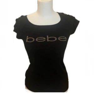 Svart kortärmad topp från Bebe - Snygg svart topp från Bebe med korta ärmar och stenar som bildar loggan framtill! 💕 Toppen har snörningsdetaljer på axlarna🙃 alla stenar är kvar och jätte bra skick! storlek S men är även väldigt stretchig 😁