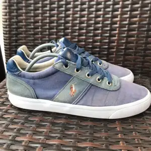 Snygga blå sneakers från Polo i textil och läder. De har en klassisk design med snörning och en vit sula. Perfekta för vardagsbruk och ger en stilren look. Märket Polo syns tydligt på sidan och hälen.