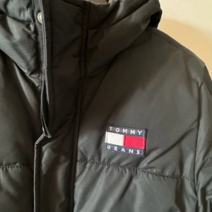 Svart pufferjacka från Tommy Jeans - Säljer en snygg svart pufferjacka från Tommy Jeans. Jättefint skick. Den har en broderad logga på bröstet och text på ärmen. Jackan är långärmad med dragkedja och knappar framtill. Perfekt för kalla dagar!