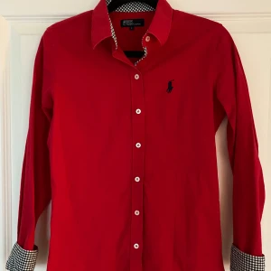 Röd skjorta från Ralph Lauren - Snygg röd skjorta från Polo Ralph Lauren i storlek S men liten i storleken. Figurnära passform. Den har långa ärmar, broderad logga på bröstet och stiliga rutiga inslag vid kragen och ärmsluten. Perfekt för både vardag och fest!