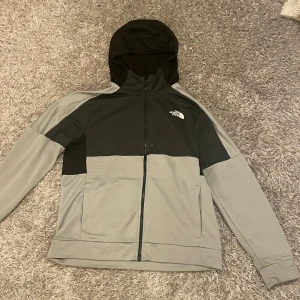 Grå och svart jacka från The North Face - Säljer en snygg grå och svart kofta i funktionsmaterial från The North Face i storlek L. Jackan har en huva och dragkedja framtill, perfekt för höst och vår. Den är i mycket bra skick och passar både till vardags och lättare utomhusaktiviteter.