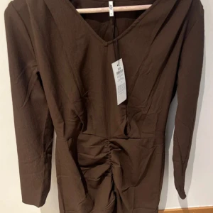 Brun långärmad klänning - Snygg brun klänning i storlek XS med lång ärm och v-ringad hals. Perfekt för höst och vinter med sin stilrena design. Klänningen har en smickrande passform och är i nytt skick med etikett kvar. Perfekt för både vardag och fest!