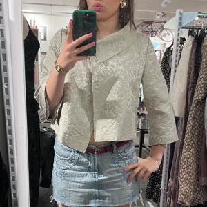 Beige brokadjacka med trekvartsärm - Säljer en superfin beige jacka i brokad med ett elegant blommönster. Jackan har trekvartsärmar och en unik krage med en stor knappdetalj framtill. Perfekt för vår och höst när du vill ha något stilrent och annorlunda. Passar både till vardags och fest!🌸