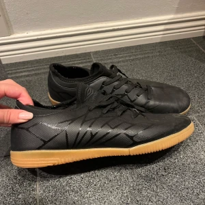 Svarta fotbollsskor med gummisula  - Säljer ett par svarta fotbollsskor i syntetmaterial med snörning. De har en cool design med mönster och en ljusbrun gummisula som ger bra grepp inomhus. Perfekta för inomhusfotboll eller futsal. Skorna är i bra skick och redo för nya matcher!