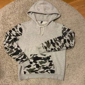 Säljer en snygg grå hoodie med coola camouflageärmar och ficka. Perfekt för en avslappnad stil och passar bra till både höst och vår. Den har en justerbar huva med snören och är i mycket bra skick. Perfekt för dig som gillar streetwear!