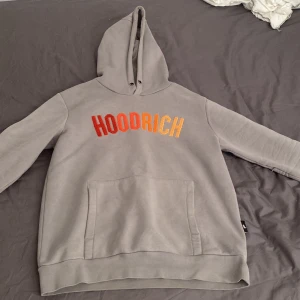 Grå hoodie från Hoodrich - Säljer en grå hoodie från Hoodrich i mycket bra skick. Den har en stor, bekväm huva och en praktisk magficka. Texten 'HOODRICH' är tryckt i en cool gradient från rött till gult. Perfekt för en avslappnad stil eller streetwear-look.