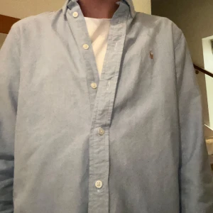 Ljusblå skjorta från Ralph Lauren - Säljer en klassisk ljusblå skjorta från Ralph Lauren i mycket bra skick. Skjortan har en normal passform och är långärmad med knappar framtill.  Passar perfekt till jeans. Passar dig som är 170-175