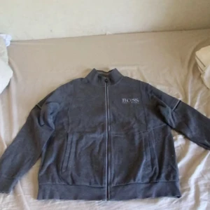 Hugo boss Kofta - Säljer en snygg grå kofta från Boss i bra skick. Den har en dragkedja framtill och långa ärmar. Perfekt för höst och vår, med en stilren design som passar till det mesta. Jackan är i en normal passform och har ribbade muddar vid ärmslut och nederkant.