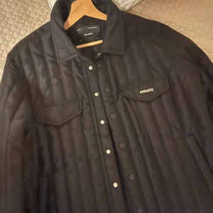 Svart quiltad jacka från Axel Arigato - Säljer en snygg svart quiltad jacka från Axel Arigato. Den har en stilren design med knappar framtill och två bröstfickor. Perfekt för höst och vår, och den är i mycket bra skick. Passar både till vardags och lite finare tillfällen.