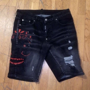 Svarta jeansshorts från Dsquared2 - Säljer ett par coola svarta jeansshorts från Dsquared2 med unika röda detaljer och slitningar. Perfekta för en avslappnad sommarlook. Shortsen har knappar och dragkedja framtill. Passar perfekt för både vardag och fest!