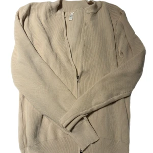 Cardigan - Säljer en stilren beige cardigan med dragkedja framtill. Den är ribbad och har långa ärmar, perfekt för både höst och vår.  Aldrig använd då den va för stor för mig skulle säga att den sitter mer som en M