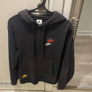 Svart hoodie från Nike - Säljer en svart hoodie från Nike i storlek S. Den har en klassisk design med färgglada Nike-loggor i rött, blått och gult på framsidan. Perfekt för en avslappnad stil och passar till alla säsonger. Den har en stor ficka fram och justerbar huva. Tar emot byten med