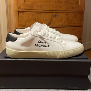 Vita sneakers från Saint Laurent - Säljer ett par snygga vita sneakers från Saint Laurent i skinn. De har en stilren design med svart häl och Saint Laurent-logga på sidan. Perfekta för både vardag och fest! Skorna har inte använts någon gång och kommer med originalförpackning. 