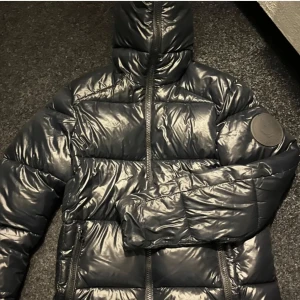 Save The Duck Jacka - Säljer en snygg svart pufferjacka i mycket bra skick. Jackan har en glansig yta och dragkedja framtill. Perfekt för kalla dagar och håller dig varm och stilren. Passar både till vardags och lite finare tillfällen.