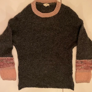 Grå stickad tröja från Levate - Mysig grå stickad tröja från Levate med rosa detaljer vid hals och ärmslut. Perfekt för kyliga dagar med sin varma och bekväma passform. Tröjan har långa ärmar och en ribbad struktur som ger en stilren look. Passar både till vardags och mysiga hemmakvällar.