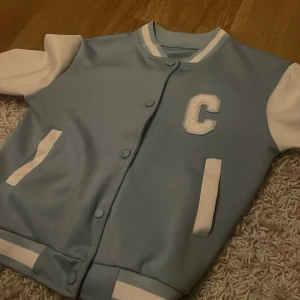 Blå och vit bomberjacka med bokstaven C - Säljer en snygg blå och vit bomberjacka med ett stort C på bröstet. Jackan har vita ärmar och blå kropp, med vita detaljer vid ärmslut och nederkant. Den stängs med knappar och har en klassisk college-stil. Perfekt för höst och vår!