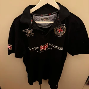 pikétröja från Geographical Norway - Säljer en snygg svart pikétröja från Geographical Norway med broderade detaljer och brittiska flaggor. Tröjan har korta ärmar och en klassisk krage. Perfekt för en avslappnad men stilren look. Passar bra till både vardag och fest! (Passar L)