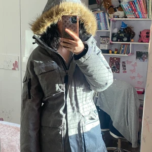 Grå parkajacka med pälskantad huva - Säljer en grå parkajacka i acceptabelt skick med en mysig pälskantad huva. Jackan har långa ärmar och praktiska fickor. Perfekt för kalla vinterdagar. Den har en dragkedja framtill och justerbara ärmslut. Defekter förekommer men jackan fungerar som vanligt och har inga hinder.