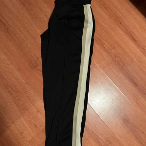 Svarta byxor med ränder från Bershka - Säljer ett par svarta byxor från Bershka i storlek M. De har ränder längs sidorna och en bekväm elastisk midja med snörning. Perfekta för en avslappnad stil eller träning. Passar bra till både vardag och sportiga aktiviteter. Hör av er vid frågor eller funderingar!