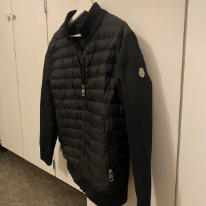 Svart quiltad jacka från Moncler - Säljer en snygg svart quiltad jacka från Moncler. Den har långa ärmar och en dragkedja framtill. Perfekt för höst och vår, med en stilren design och Moncler-logga på ärmen. Jackan är i mycket bra skick och passar både till vardags och lite finare tillfällen.