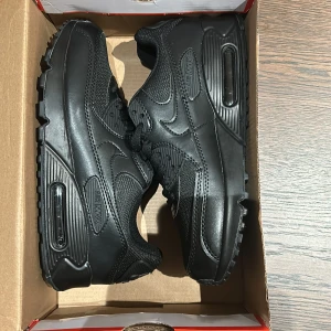 Svarta Nike Air Max i syntet - Säljer ett par svarta Nike Air Max i syntetmaterial. De är i nytt skick och har en klassisk design med snörning och den ikoniska Air Max-sulan. Perfekta för både vardag och träning. Storlek är inte specificerad, men de ser ut att vara i standard herrstorlek. Skorna är helt svarta vilket gör dem lätta att matcha med olika outfits.