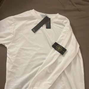 Vit långärmad tröja från Stone Island - Säljer en snygg vit långärmad tröja från Stone Island. Den är i nytt skick med etiketter kvar. Perfekt för en stilren look och passar till alla tillfällen. Tröjan har en klassisk design med Stone Island-loggan på ärmen.