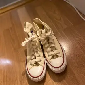 Säljer ett par klassiska höga Converse sneakers i beige med snörning. De har den ikoniska röda och blå loggan på sidan och en vit sula. Perfekta för en avslappnad stil och passar till nästan allt. Använda men i bra skick!