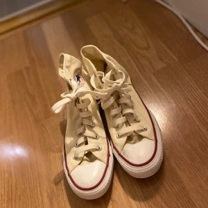 Beige höga sneakers från Converse - Säljer ett par klassiska höga Converse sneakers i beige med snörning. De har den ikoniska röda och blå loggan på sidan och en vit sula. Perfekta för en avslappnad stil och passar till nästan allt. Använda men i bra skick!