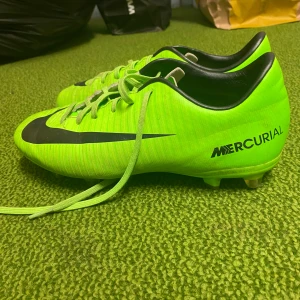 Nike Mercurial fotbollsskor - Säljer ett par grymma Nike Mercurial fotbollsskor i neonlimegrön färg med svarta detaljer. Skorna har snörning och är perfekta för fotbollsträning eller match. De är i bra skick och redo för nya äventyr på planen. Passar dig som vill sticka ut med stil och prestanda!