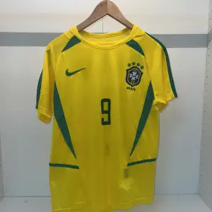 Säljer en gul Brasilien fotbollströja från Nike i nytt skick. Tröjan har korta ärmar och är prydd med gröna detaljer samt nummer 9 på framsidan och baksidan. Den har också namnet 'Ronaldo' på ryggen. Tillverkad med Nike Breathe-teknologi för att hålla dig torr och bekväm. Perfekt för fotbollsfans! Storlek s/m priset kan diskuteras!🏆