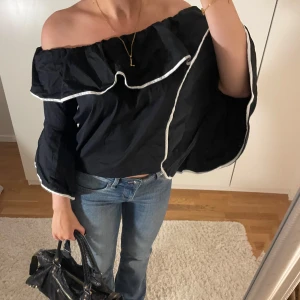 Svart offshoulder topp med volang - Säljer en superfin svart offshoulder topp med volangdetaljer. Den har långa ärmar och en lös passform, perfekt för både vardag och fest. Toppen är i mycket bra skick och har vita kantdetaljer som ger en snygg kontrast. Passar perfekt till jeans eller kjol!