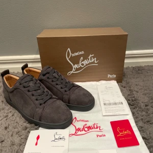 Grå mockasneakers från Christian Louboutin - Säljer ett par snygga grå sneakers i mocka från Christian Louboutin. De har den ikoniska röda sulan och klassisk snörning. Perfekta för både vardag och fest! Skorna är i nytt skick och kommer med originalförpackning och tillbehör.