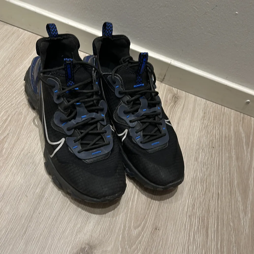 Säljer ett par svarta Nike vision react sneakers i mycket bra skick. De har snygga blå detaljer och vit snörning. Perfekta för både träning och vardag. Skorna har en modern design med bekväm passform och är gjorda i syntetmaterial.. Skor.