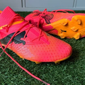 Rosa fotbollsskor från Puma - Säljer ett par grymma rosa fotbollsskor från Puma i syntetmaterial. De har en cool svart detalj på sidan och orangea dobbar för bästa grepp på planen. Perfekta för dig som vill sticka ut på fotbollsplanen! Skorna har snörning för en tight passform.