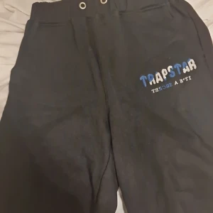 Svarta mjukisbyxor från Trapstar - Säljer ett par svarta mjukisbyxor från Trapstar i bra skick. De har en elastisk midja och en cool logga i blått och vitt på sidan. Perfekta för en avslappnad stil eller chill dagar hemma. Passar bra till sneakers och en hoodie! ( ai )