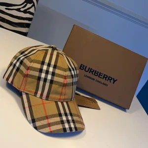 Rutig keps från Burberry - Säljer en snygg  keps från Burberry i klassiskt mönster. Kepsen är helt ny med alla tags kvar. Perfekt för att ge din outfit en lyxig touch. Kommer med originalförpackning och etikett.