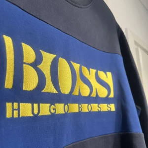 Blå och svart tröja från Hugo Boss - Säljer en snygg blå och svart tröja från Hugo Boss med gul logga på bröstet. Tröjan är långärmad och perfekt för både höst och vår. Den har en normal passform och är i mycket bra skick. Perfekt för en casual look eller när du vill känna dig extra stilren.