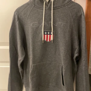 Grå hoodie från GANT - Säljer en snygg grå hoodie från GANT i mycket bra skick. Den har en broderad logga och en stor ficka framtill. Perfekt för höst och vinter, och den är supermjuk och bekväm. Passar både till vardags och chillkvällar. 🧢