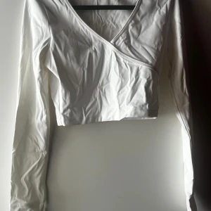Vit omlottopp från MOT - Säljer en stilren vit omlottopp från MOTF i storlek S. Toppen har en v-ringad design och är tillverkad i ett mjukt och stretchigt material av elastan och polyamid. Perfekt för både vardag och fest!