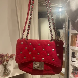 Röd axelväska med nitar - Säljer en supercool röd axelväska i mocka med silverfärgade nitar. Väskan har en kedjerem som ger en edgy look och ett snyggt lås framtill. Perfekt för att piffa upp vilken outfit som helst! 👜✨